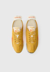 Mustársárga Reebok sportcipők, tónusos burkolatokkal, narancssárga részletekkel, lapos fűzőkkel, valamint puha, párnázott nyelvvel és gallérral.