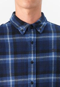 Blue plaid flannel shirt featuring a button-up collar, black buttons, and white accents, with a soft texture and checkered pattern.