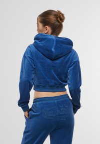 Felpa corta blu in morbido velluto con cappuccio, presenta polsini e orlo a costine. Logo "Von Dutch" ricamato sui pantaloni abbinati.