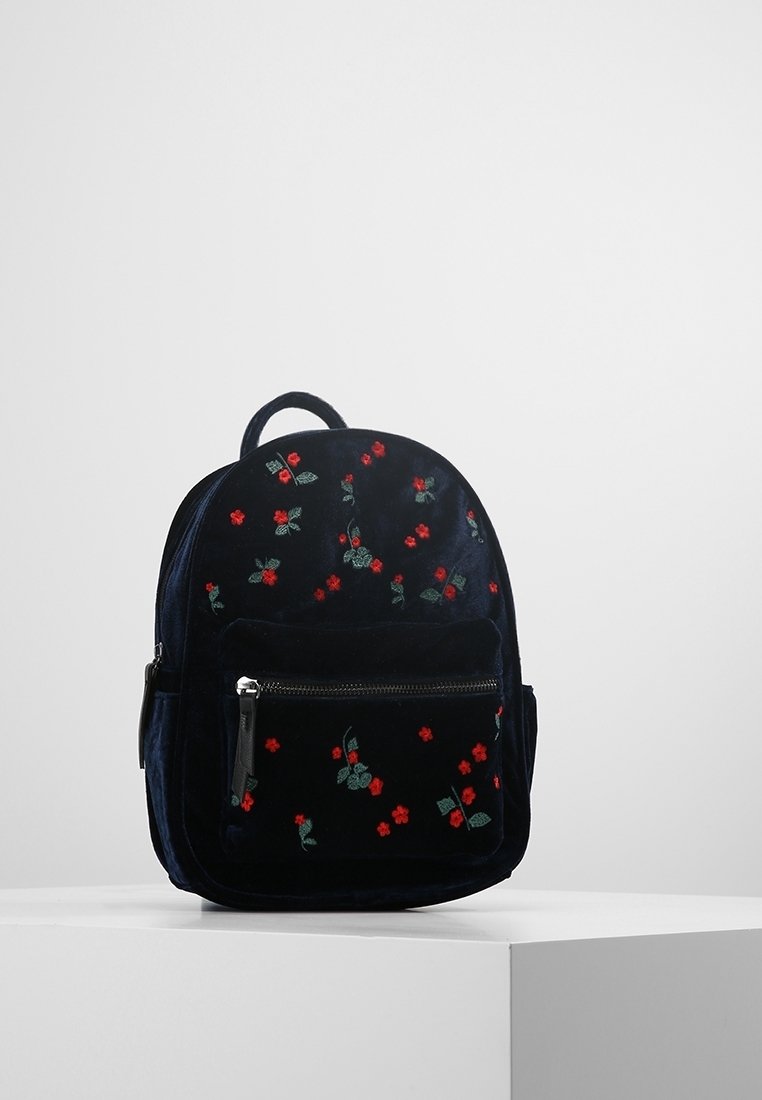 Petit sac à dos noir avec une poche zippée à l'avant, décoré de broderies de fleurs rouges et de feuilles vertes, posé sur un piédestal blanc.