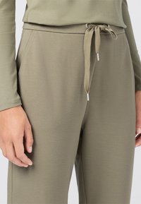 Olivgrüne, weiche Jogginghosen aus Stoff mit elastischem Bund, verstellbarem Kordelzug und silbernen Metallspitzen. Nahaufnahme von Hand- und Beindetail.