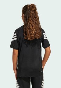 Črna atletska majica s kratkimi rokavi, ki ima tri bele črte na vsakem rami in zasnovo sproščenega kroja. Gladko, lahko blago.