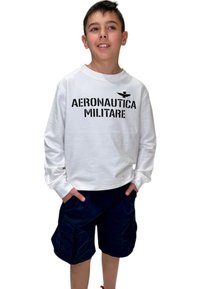 Camicia bianca a maniche lunghe con "AERONAUTICA MILITARE" in lettere nere in grassetto; abbinata a pantaloni cargo blu. Il bambino è in piedi con le mani nelle tasche.
