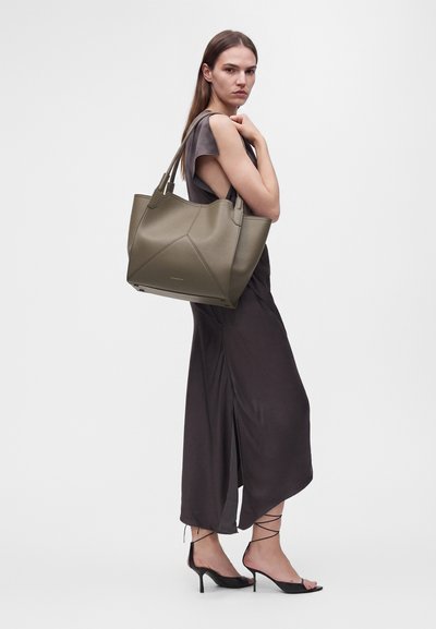 Femme en robe sombre sans manches et talons noirs portant un grand sac cabas vert olive sur l'épaule devant un fond uni.