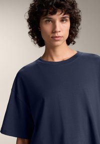 Tricou din bumbac de culoare bleumarin, cu o croială relaxată, gât rotund și mâneci scurte. Textură moale, design minimalist, fără modele sau accente vizibile.