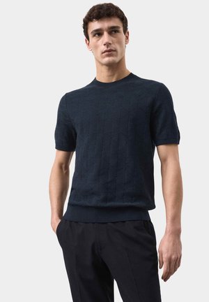 Jonge man die een donkerblauw gebreid kortemouwen trui en zwarte broek draagt, staand met één hand in de zak tegen een effen achtergrond.
