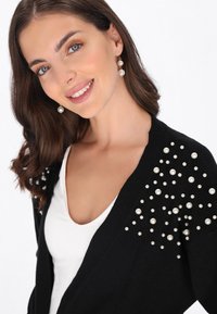 Czarny kardigan z ryflem, ozdobiony grupami perłowych akcentów na ramionach. Noszony na białej bluzce.
