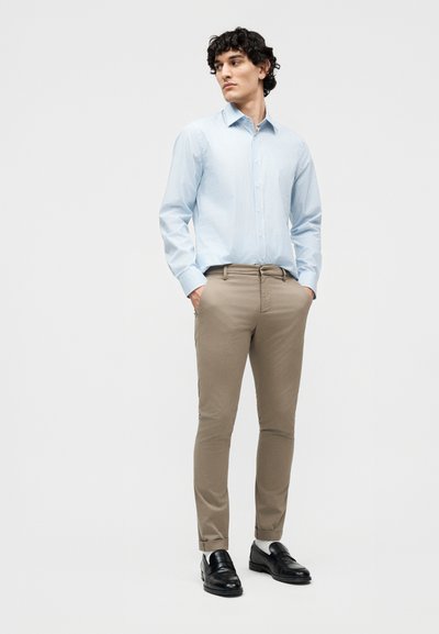 Άνδρας που φοράει ανοιχτό μπλε ριγέ πουκάμισο, μπεζ chinos με γυρίσματα, μαύρα loafers, στέκεται με τα χέρια στις τσέπες, κοιτάζοντας προς τα αριστερά.