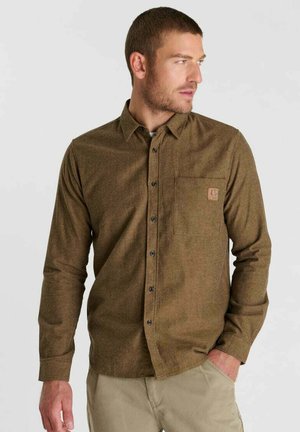 Man wearing brown button-up shirt and beige pants looking to his right with one hand in his pocket against plain background.