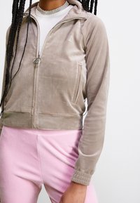Beige kort zip-up jacka i mjuk sammet med en framlapp och en logodetalj. Matchad med ljusrosa slim fit-byxor.