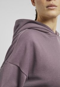Bluza z kapturem w stonowanym fioletowym kolorze, wykonana z miękkiego materiału. Posiada luźny krój, opadające ramiona oraz dużą, usztywnioną kieszeń w kapturze.