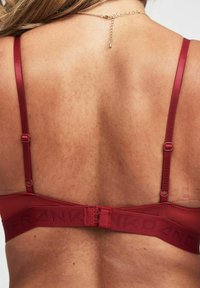 Red fabric bra with thin straps, adjustable closures, and a band featuring a logo in a subtle pattern. Smooth texture and minimal design.