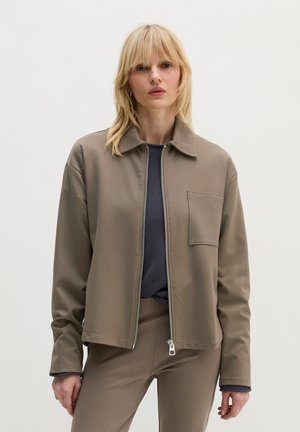 Vrouw met blond haar, gekleed in een taupekleurig jack met rits en borstzak, over een donker shirt en bijpassende taupekleurige broek, staand tegen een effen achtergrond.