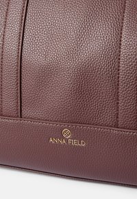 Mala de mão em imitação de pele texturizada bordeaux, com detalhes cosidos e um logótipo dourado "ANNA FIELD" no painel frontal inferior.