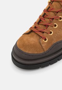 Stivale da trekking in suede marrone con lacci intrecciati rossi e marroni, occhielli dorati e una spessa suola in gomma nera. Punta arrotondata e dettagli cuciti.