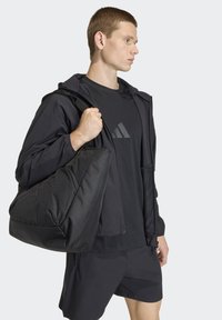 Czarny strój sportowy obejmujący lekką kurtkę, koszulkę z emblematem oraz szorty. Model trzyma dużą czarną torbę z tkaniny.