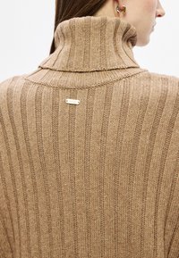 Μπεζ πλεκτό πουλόβερ με ψηλό γιακά, με κάθετη ρίγα και πλακέτα επωνυμίας σε χρυσό τόνο στο πίσω μέρος. Απαλή υφή.