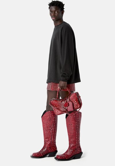 Jeune personne portant un sweat-shirt noir oversize, une minijupe rouge à motif crocodile et des bottes hautes assorties, tenant un sac à main rouge texturé.