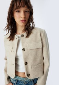 Chaqueta corta beige hecha de una tela suave, con un escote redondo, dos bolsillos frontales y tres botones marrones como acentos de hardware.