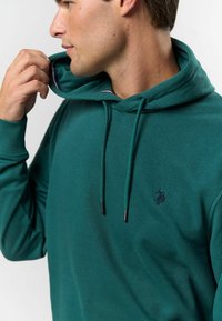 Zöld kapucnis pulóver puha anyagból, húzózsinórral ellátott kapucnival, tónusos logóval és kontrasztos belső kapucni szegéllyel. Sima textúra, laza fazon.