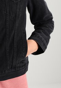 Chaqueta de denim negra con un bolsillo frontal, costuras visibles y un dobladillo acanalado. La manga tiene un detalle con puño, combinado con una tela rosa en el interior.