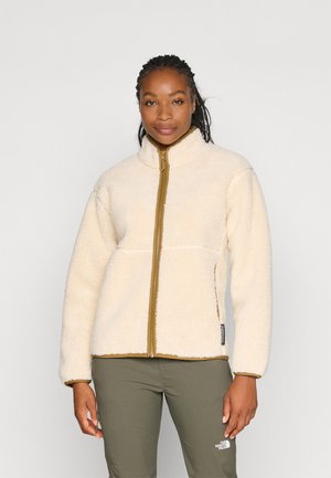 Vrouw draagt een crèmekleurige fleecejas met ritssluiting aan de voorkant en olijfgroene broek, staande tegen een effen lichte achtergrond.