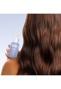 Coco & Eve Haarserum in einer klaren Glasflasche mit einem weißen Tropferdeckel. Langes, gewelltes, braunes Haar ist im Hintergrund sichtbar.