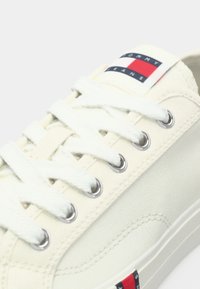 Λευκό πάνινο αθλητικό παπούτσι με στρογγυλές ασημί τρύπες και λευκά κορδόνια. Διαθέτει ετικέτα με το λογότυπο Tommy Jeans σε κόκκινο, λευκό και μπλε στη γλώσσα του παπουτσιού.