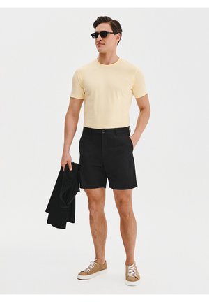 Czarne szorty i jasnoniebieska koszulka. Model nosi okulary przeciwsłoneczne, trzyma czarną kurtkę i ma na sobie beżowe sportowe buty z białymi podeszwami.