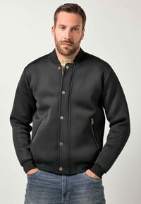 Giacca bomber nera con una texture liscia, caratterizzata da un colletto a coste, bottoni a pressione e tasche laterali con cerniera. Indossata sopra un maglione beige.