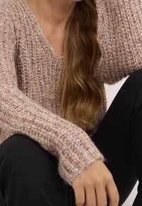 Pull en tricot légèrement texturé, rose et beige, avec une coupe ample, des manches longues et des poignets côtelés, porté sur un pantalon noir.