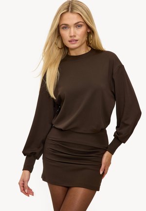 Femme blonde portant une mini-robe moulante à manches longues de couleur brun foncé et de grandes boucles d'oreilles en or, debout avec une main sur la hanche.