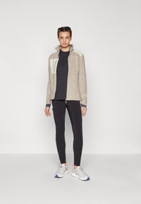 Beige fleecová bunda na zip s vysokým límcem, která má bílou část a zipovou kapsu. Kombinována s tmavým topem a černými legínami, doplněná šedými teniskami.