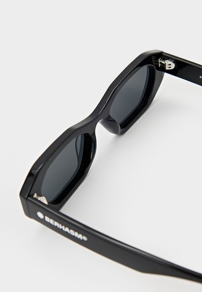 Black sunglasses with a thick, angular frame, featuring dark lenses and metallic accents on the hinges. Branding visible on the temples.
