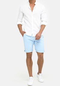 Camisa blanca de lino con botones, pantalones cortos azul claro arremangados y zapatillas blancas. Enfocado en tejidos transpirables y un ajuste casual y a medida.