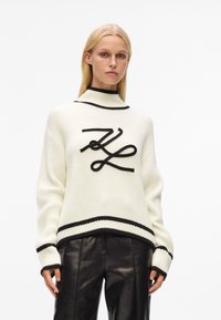 Pull en maille blanc à rayures noires, col haut et détail de logo noir bien visible sur le devant. Porté avec un pantalon en cuir noir.