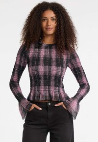 Frau mit lockigem Haar, die ein langärmliges Oberteil mit pink-schwarzem Karomuster und ausgestellt Bündchen sowie schwarze Hosen trägt, Hände in den Taschen, neutraler Hintergrund.