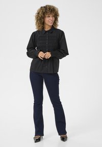 Camicia nera con bottoni, dettagli arricciati sul petto e polsini elasticizzati, abbinata a jeans a zampa d'elefante blu scuro e decolletè neri in vernice.