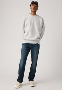 Svetlosivá mikina z mäkkej látky s voľným strihom, kombinovaná s tmavomodrými džínsami. Model má na hlave čiernu čiapku a na nohách biele tenisky.