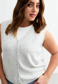Grijze mouwloze gebreide vest met een v-hals, voorzien van kleine knopen aan de voorkant en een zachte, gestructureerde afwerking, gedragen over een witte top.