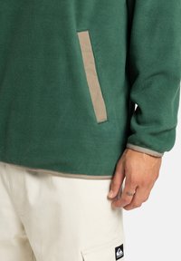 Pullover in felpa verde con una texture morbida, caratterizzato da una tasca in contrasto color tan e semplici dettagli sull'orlo, abbinato a pantaloni cargo color bianco sporco.