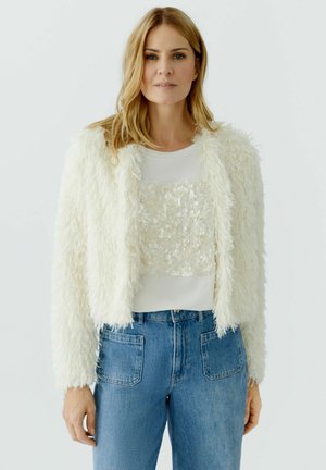 Femme aux cheveux blonds portant une veste blanche duveteuse texturée par-dessus un haut blanc à motifs floraux et un jean bleu taille haute.