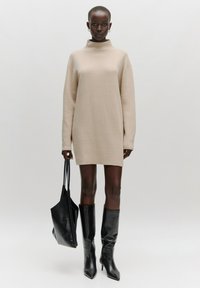 Beige trui jurk met een hoge hals, lange mouwen en een relaxte pasvorm. Gepaarde met zwarte kniehoge stiletto laarzen en een zwarte tote tas.