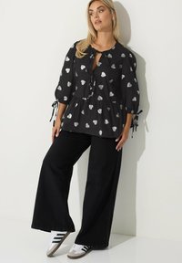 Blusa nera con stampa di cuori argentati, maniche a sbuffo e chiusura con fiocco al collo, abbinata a jeans neri a gamba larga e sneakers bianche con dettagli grigi.