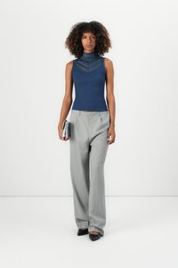 Top sem mangas em malha de cor azul-marinho com detalhes em renda, combinado com calças largas cinza com risca de giz, segurando uma carteira prateada. Sapatos planos pretos.