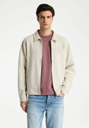 Junger Mann mit dunklen Haaren trägt eine hellbeige Zip-up-Jacke über einem mauvefarbenen Hemd und hellblauen Jeans, steht vor einem schlichten Hintergrund.