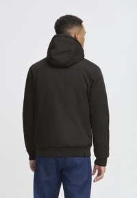 Schwarze Daunenjacke mit Kapuze, glattes Material, elastische Bündchen und Saum sowie Rückenpasse. Getragen mit blauen Jeans.