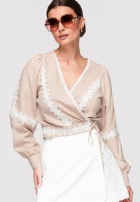 Femme portant un chemisier beige portefeuille avec bordure en dentelle blanche, jupe blanche et grandes lunettes de soleil marron sur fond gris.