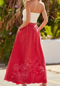 Jupe maxi en tissu rouge, ornée de motifs brodés blancs représentant des plantes, un œil et un singe. Taille élastique, silhouette fluide.