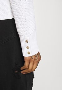 Pull blanc avec un motif texturé, poignets côtelés ornés de trois boutons métalliques, associé à un tissu en jean noir déchiré.